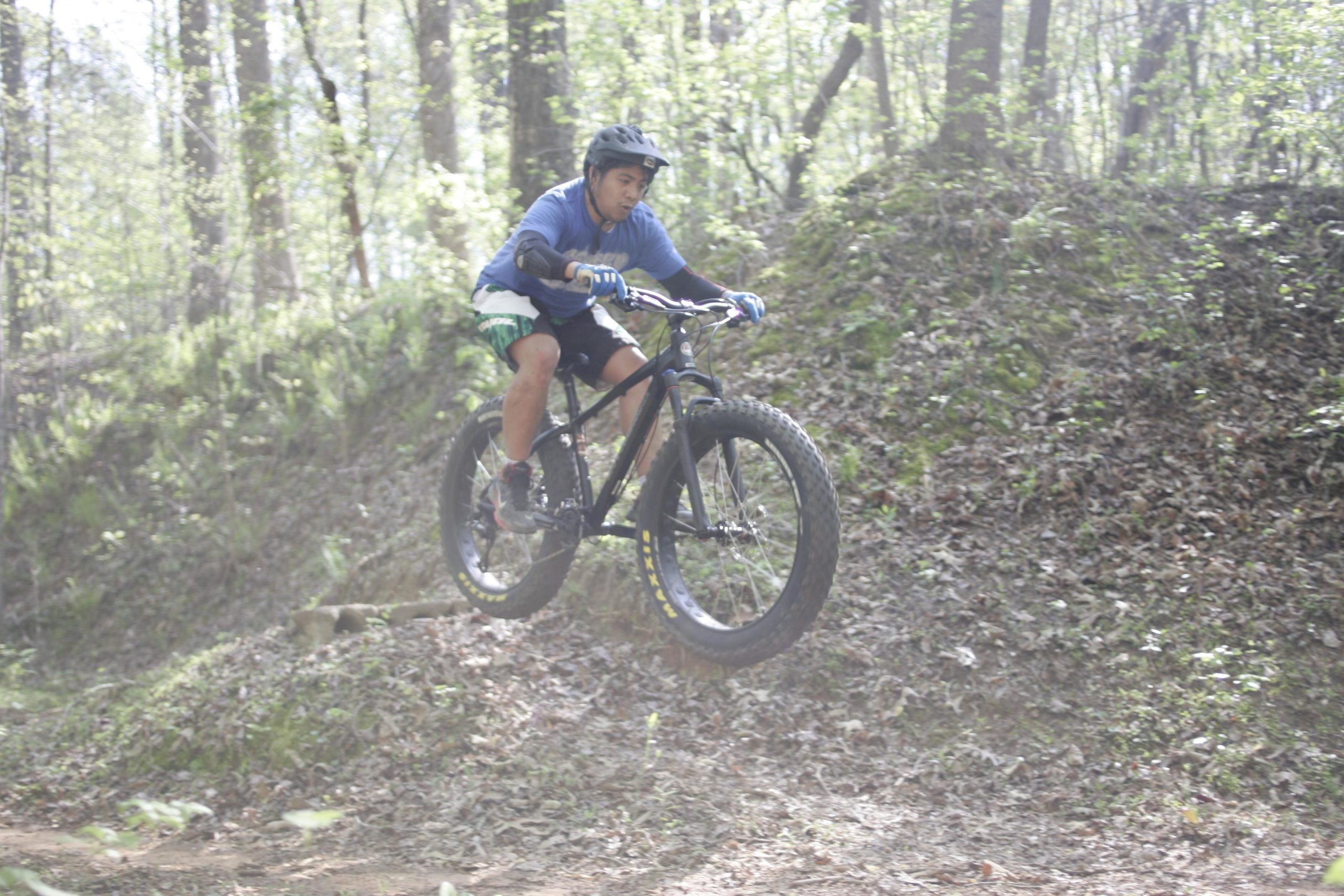 A young male cyclist in a blue shirt and shorts performs a bicycle jump on a dirt trail surrounded by trees. The cyclist is airborne, with a focused expression, demonstrating skill and agility as he navigates the natural terrain. Dust is kicked up from the trail, adding a sense of action to the scene. Salem Lake mountain bike trail.