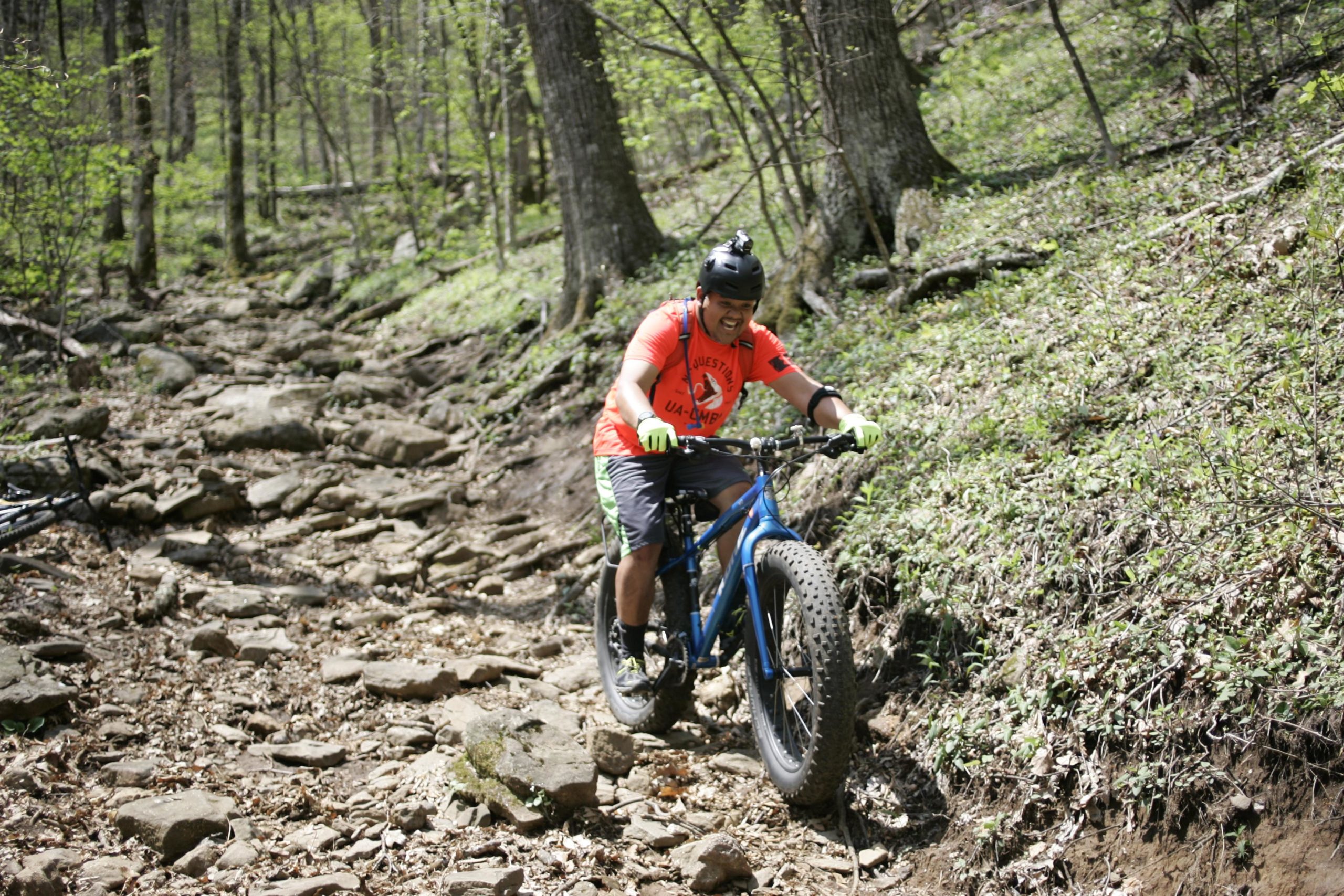 A person riding a blue fat bike on a rocky trail in a forested area. The individual is wearing a bright orange shirt, black shorts, a helmet, and is smiling as they navigate the uneven terrain surrounded by trees and greenery. Farlow Gap mountain bike trail.