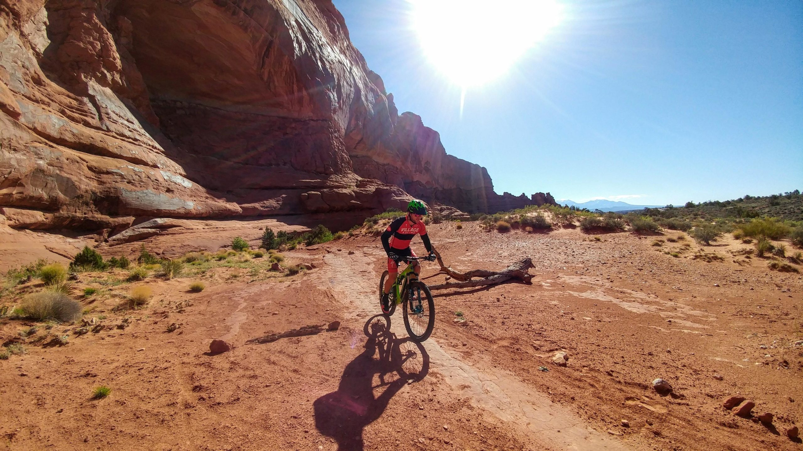 A mountain biker rides on a dusty trail surrounded by red rock formations under a bright sun. The landscape features sandy soil with sparse vegetation and a clear blue sky. Navajo Rocks mountain bike trail.