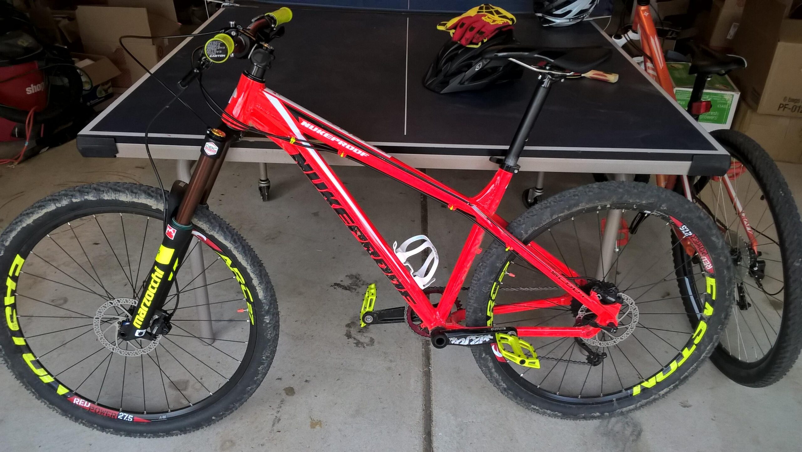Nukeproof Scout: A bright red mountain bike with yellow accents is positioned in a garage. The bike features thick tires, a sturdy frame, and visible components such as the handlebars and pedals. In the background, a ping pong table and other biking gear can be seen, indicating a casual recreational space.
