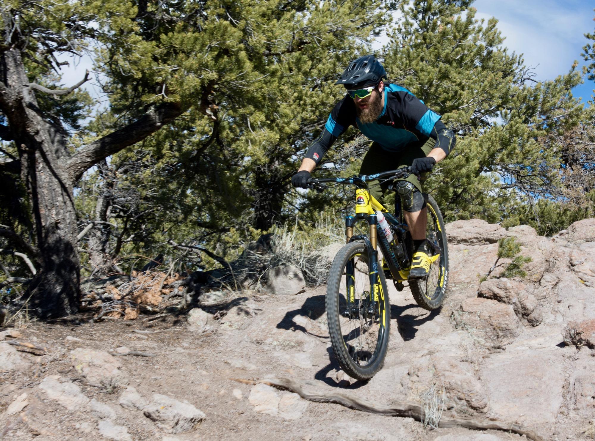 A mountain biker with a beard rides down a rocky trail in a forested area. He wears a black helmet, sunglasses, and a blue and black jersey, along with yellow shorts and gloves. The bike is yellow with black and blue accents, and the background features pine trees and rugged terrain under a clear blue sky. Unkle Nazty mountain bike trail.