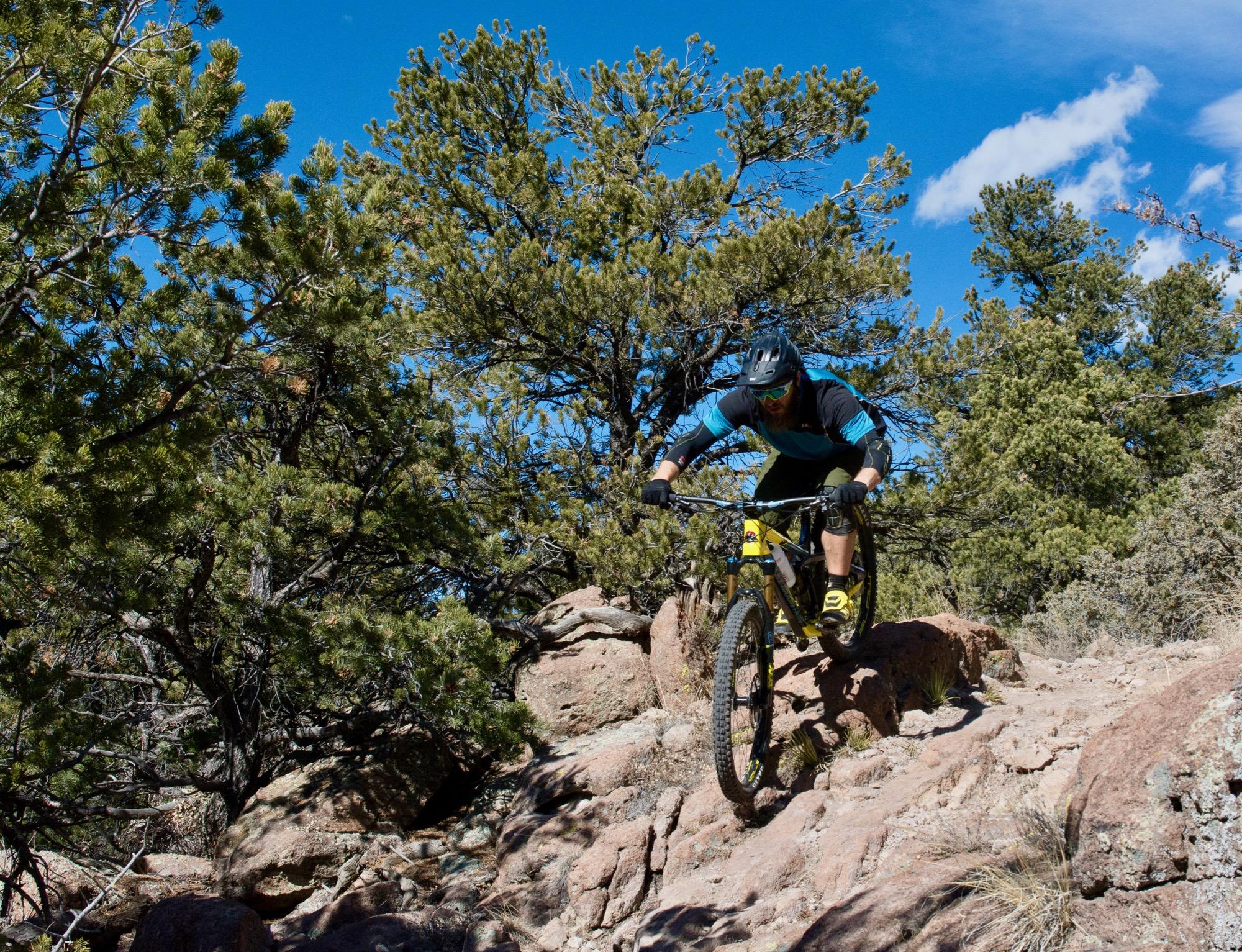 A mountain biker navigating rocky terrain in a forested area, airborne off a ledge, with a clear blue sky and scattered clouds in the background. Unkle Nazty mountain bike trail.