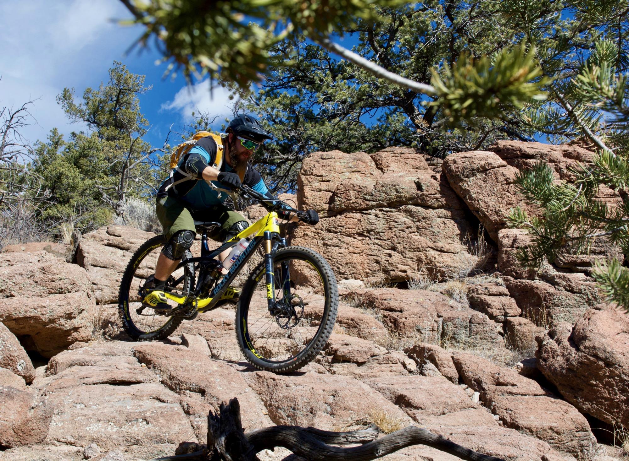 A mountain biker navigating rocky terrain, wearing a helmet and protective gear, with a backpack and sunglasses. Surrounding the biker are trees and clear blue skies, capturing the essence of outdoor adventure and cycling. Unkle Nazty mountain bike trail.
