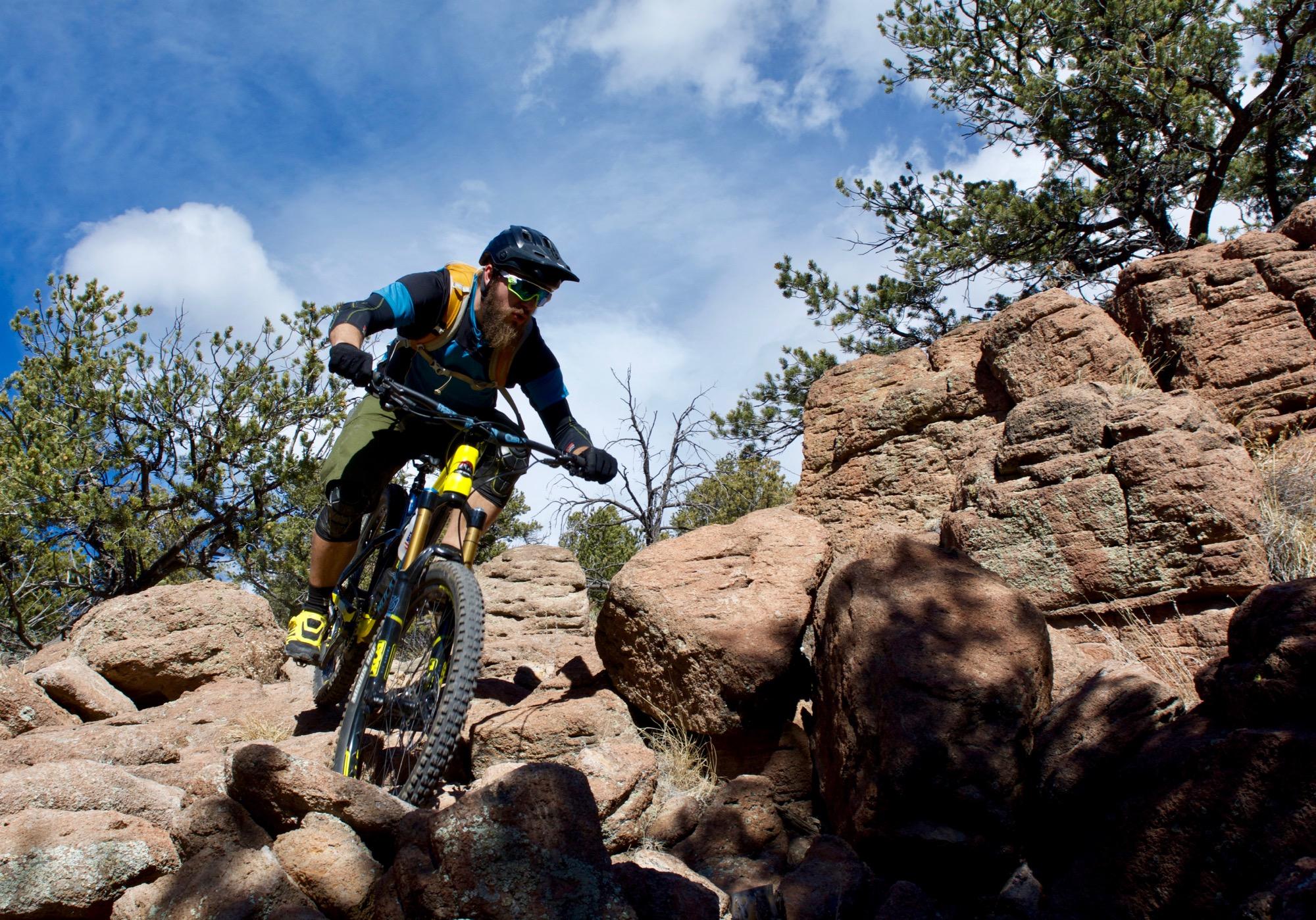 A mountain biker navigating rocky terrain under a blue sky with scattered clouds, surrounded by trees. The rider is wearing a helmet and protective gear, focused on maneuvering over boulders. Unkle Nazty mountain bike trail.