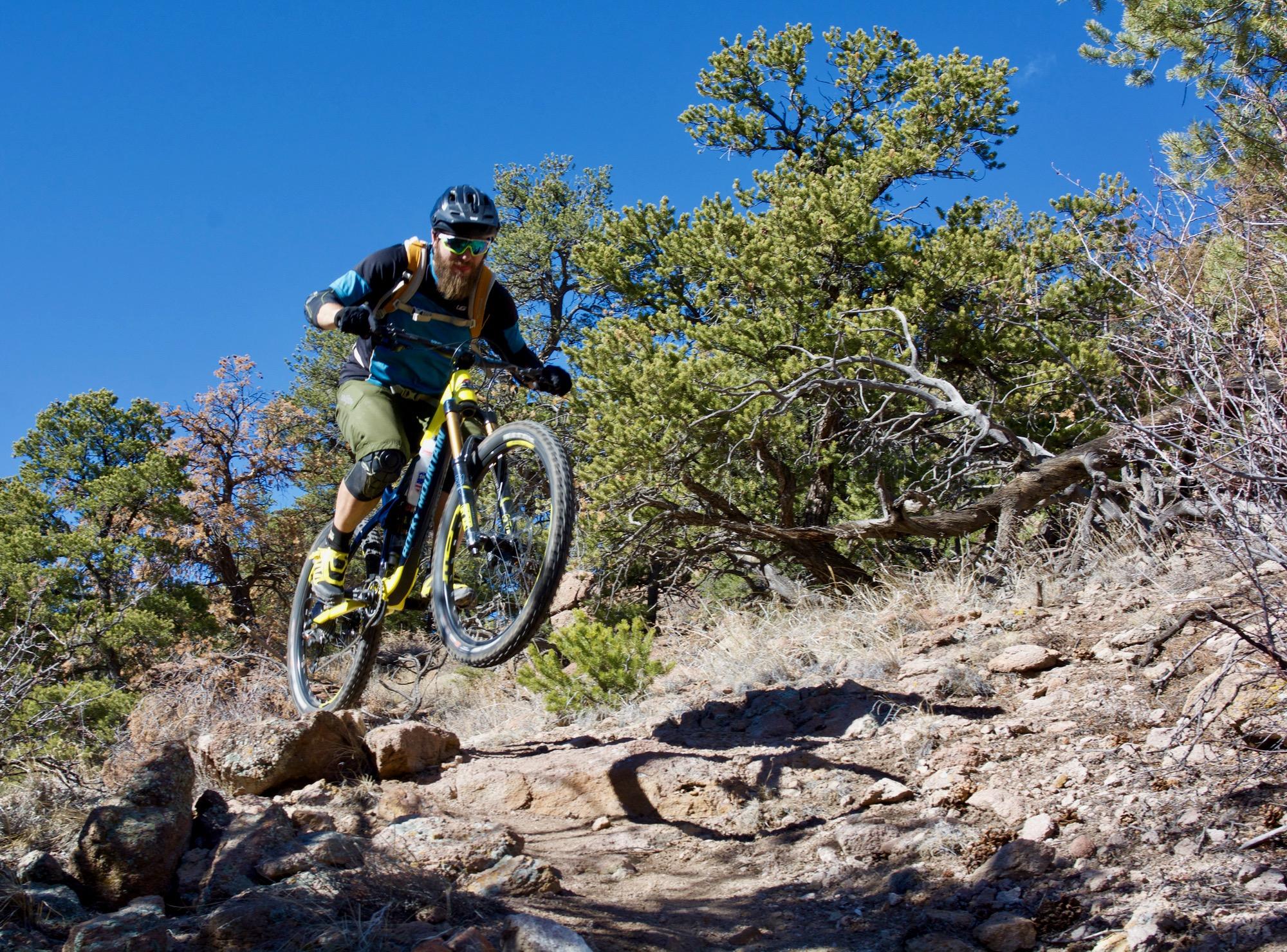 A mountain biker in a blue and black jersey and green shorts jumps over rocky terrain on a trail surrounded by trees under a clear blue sky. Unkle Nazty mountain bike trail.
