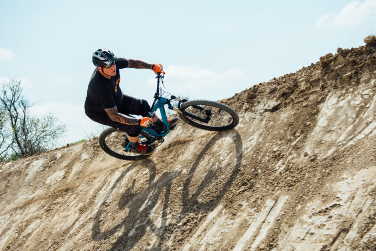 A person riding a mountain bike performs a sharp turn on a dirt hillside, showcasing skill and agility. The rider is wearing a helmet and gloves, with visible tattoos on their arms, and is positioned against a clear blue sky. Dust is kicked up from the bike tires, emphasizing movement and action.