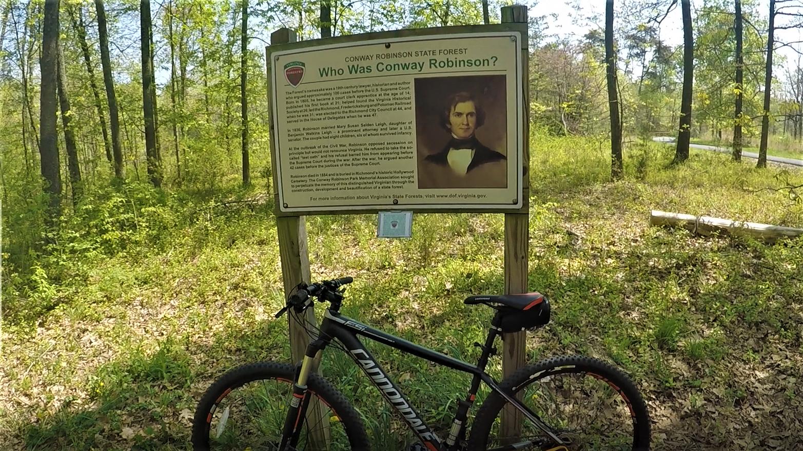A mountain bike is parked in front of an educational sign in Conway Robinson State Forest, surrounded by greenery and trees. The sign features information about Conway Robinson, including a portrait and a brief historical description of his contributions as a 19th-century lawyer, historian, and author. Conway Robinson State Forest mountain bike trail.