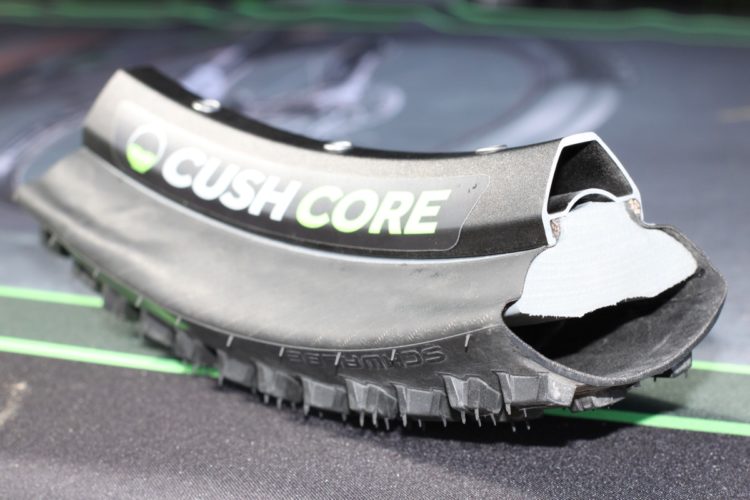 A close-up view of a cross-section of a Cush Core tire insert. The insert is black with the "Cush Core" logo displayed prominently. The inner portion shows a foam-like texture, highlighting its construction and design for improved shock absorption in tires. The background features a textured surface with green accents.