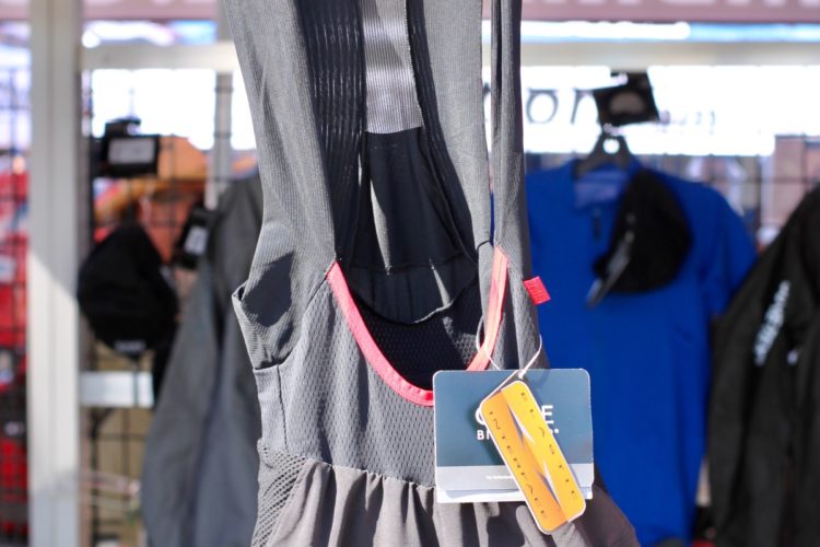 A pair of black cycling bib shorts displayed on a hanger, featuring a red inner lining and mesh panels for breathability. A price tag is attached, with various other clothing items visible in the background under a market tent.