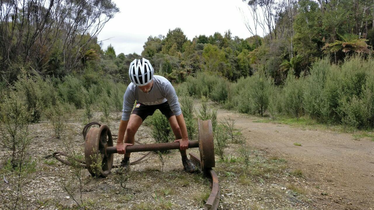 A person wearing a helmet is lifting a large, rusty piece of metal in a wooded area with sparse vegetation. The background features trees and a dirt path, suggesting an outdoor exercise or training environment. The Timber Trail mountain bike trail.
