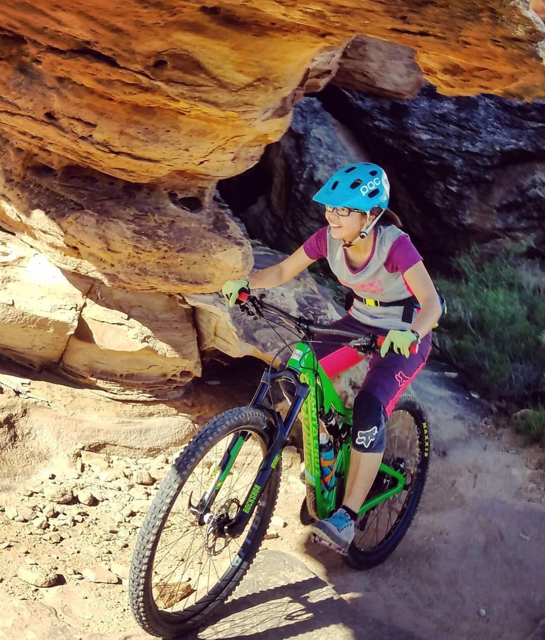 A person with glasses riding a green mountain bike on a rocky trail, smiling as they navigate through a narrow passageway formed by large boulders. The scene is set in a natural outdoor environment with scattered greenery and sunlight illuminating the rocks. The rider is wearing a blue helmet and appropriate biking gear. Zen Trail mountain bike trail.