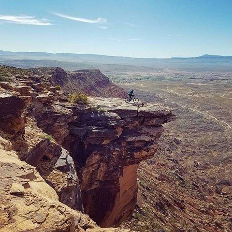 A mountain biker navigating along the edge of a rocky cliff with a vast landscape visible in the background. The scene showcases an arid environment with distant mountains and clear blue skies. Zen Trail mountain bike trail.