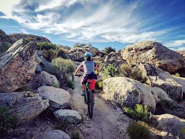 A person riding a mountain bike on a rocky trail surrounded by boulders and sparse vegetation, with a clear blue sky and wispy clouds above. Zen Trail mountain bike trail.