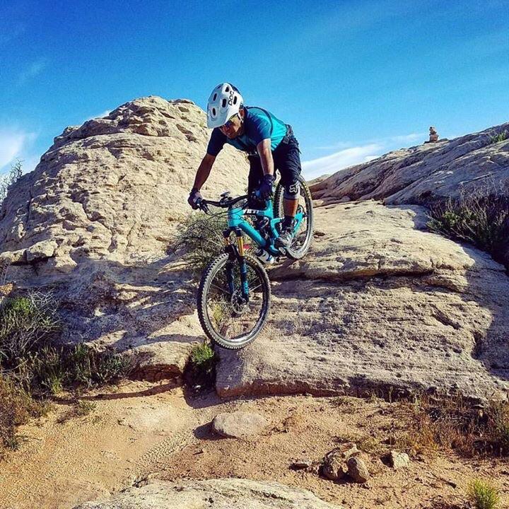 A mountain biker in a blue and black outfit maneuvers over rocky terrain under a clear blue sky, showcasing agility and skill as he rides a teal mountain bike. The landscape features rugged rocks and sparse vegetation, typical of a natural outdoor biking trail. Zen Trail mountain bike trail.