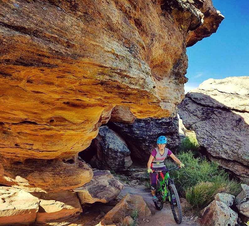 A mountain biker navigating a rocky trail under a large overhang, surrounded by golden-hued rocks and blue sky. The cyclist is wearing a blue helmet and colorful biking gear, with greenery visible along the path. Zen Trail mountain bike trail.