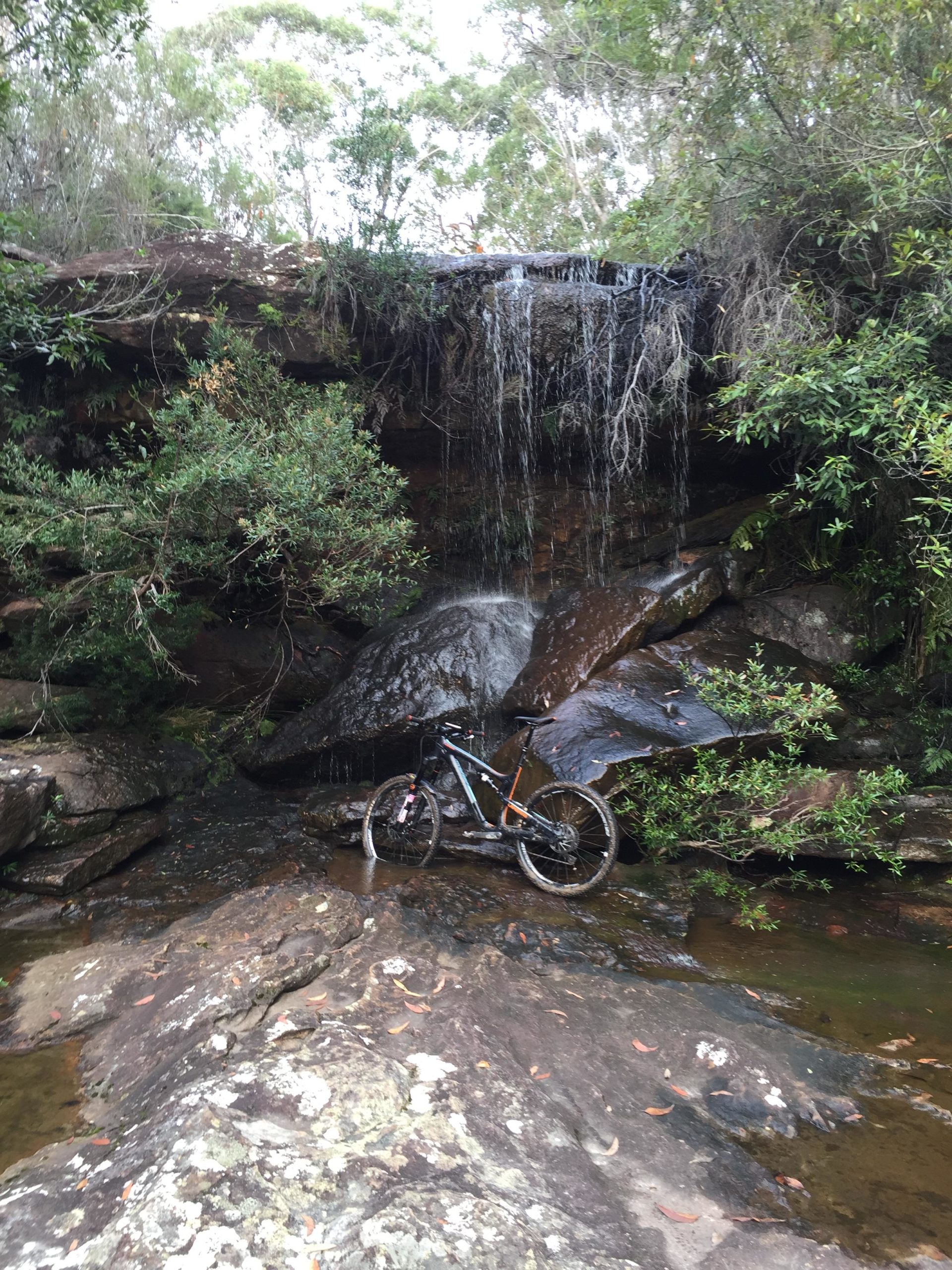 A mountain bike resting on rocky terrain beside a small waterfall, surrounded by lush greenery and trees. Deep creek reserve mountain bike trail.