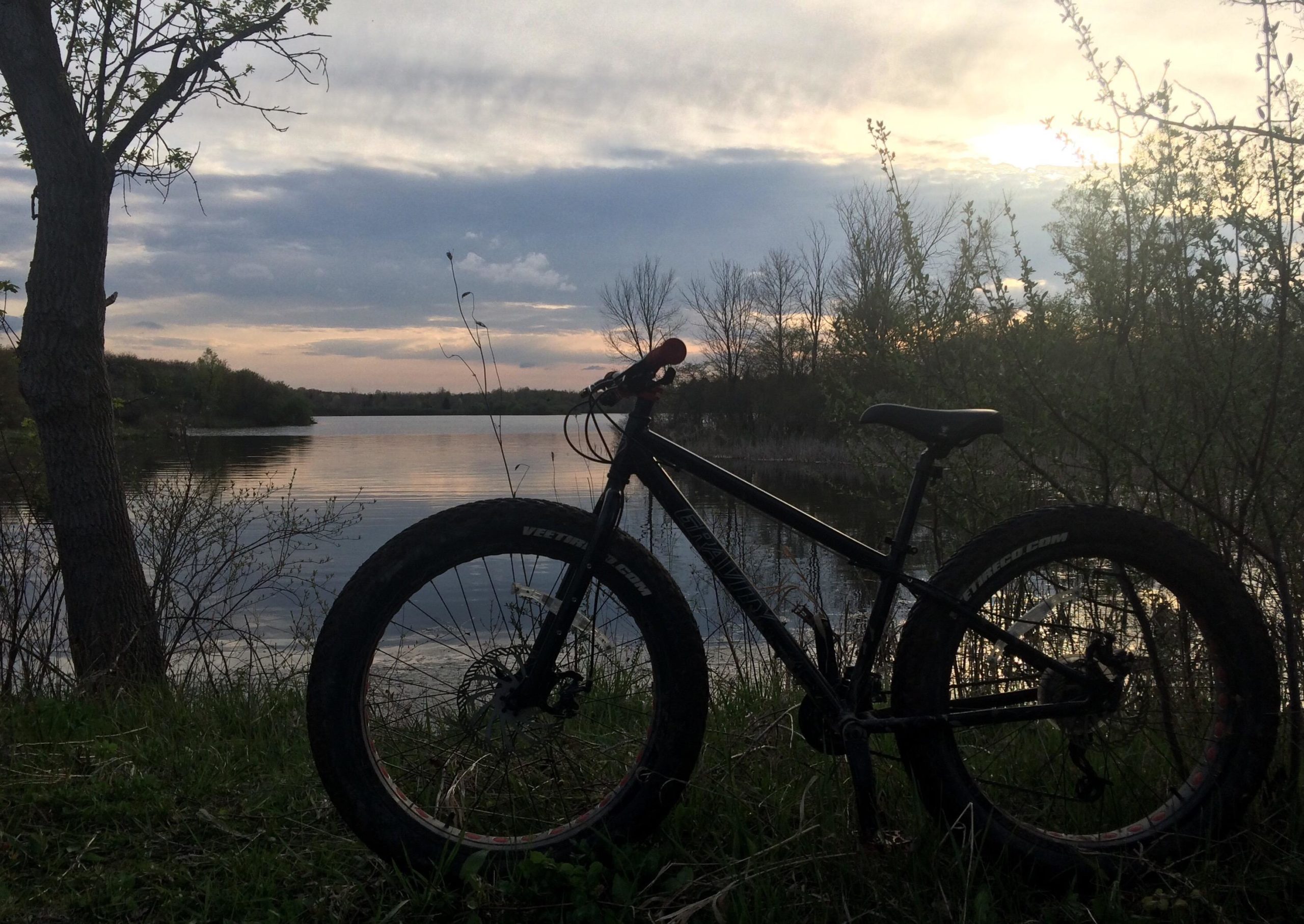 A black fat tire mountain bike stands on the grassy bank beside a serene lake at sunset, with calm waters reflecting the soft pastel colors of the sky. Bare trees and gentle vegetation frame the scene, creating a tranquil atmosphere. Sleepy Hollow mountain bike trail.