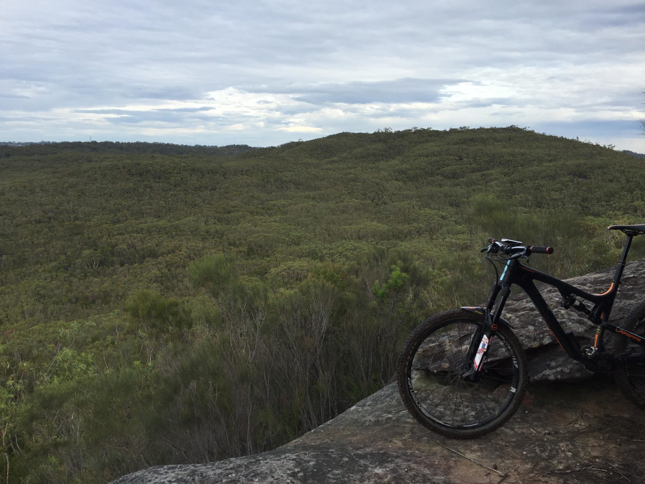 A mountain bike resting on a rock outcrop overlooking a lush green landscape with rolling hills under a cloudy sky. Deep creek reserve mountain bike trail.