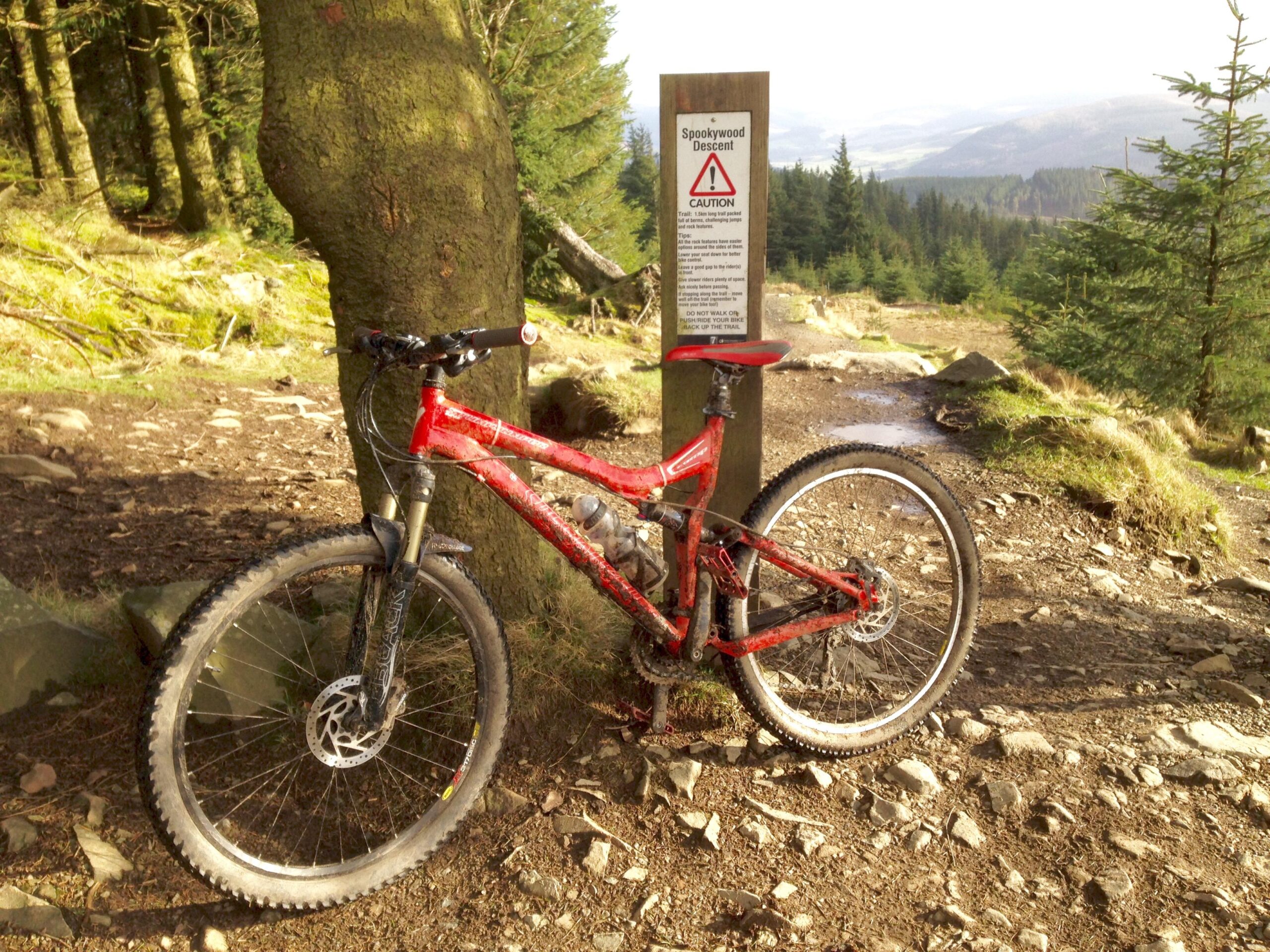 Specialized Stumpjumper FSR: A red mountain bike leaning against a tree near a trail sign that reads "Spookywood Descent," indicating caution with warnings for bikers. The background features a wooded area with gravel and rocks, and a scenic view of hills in the distance.