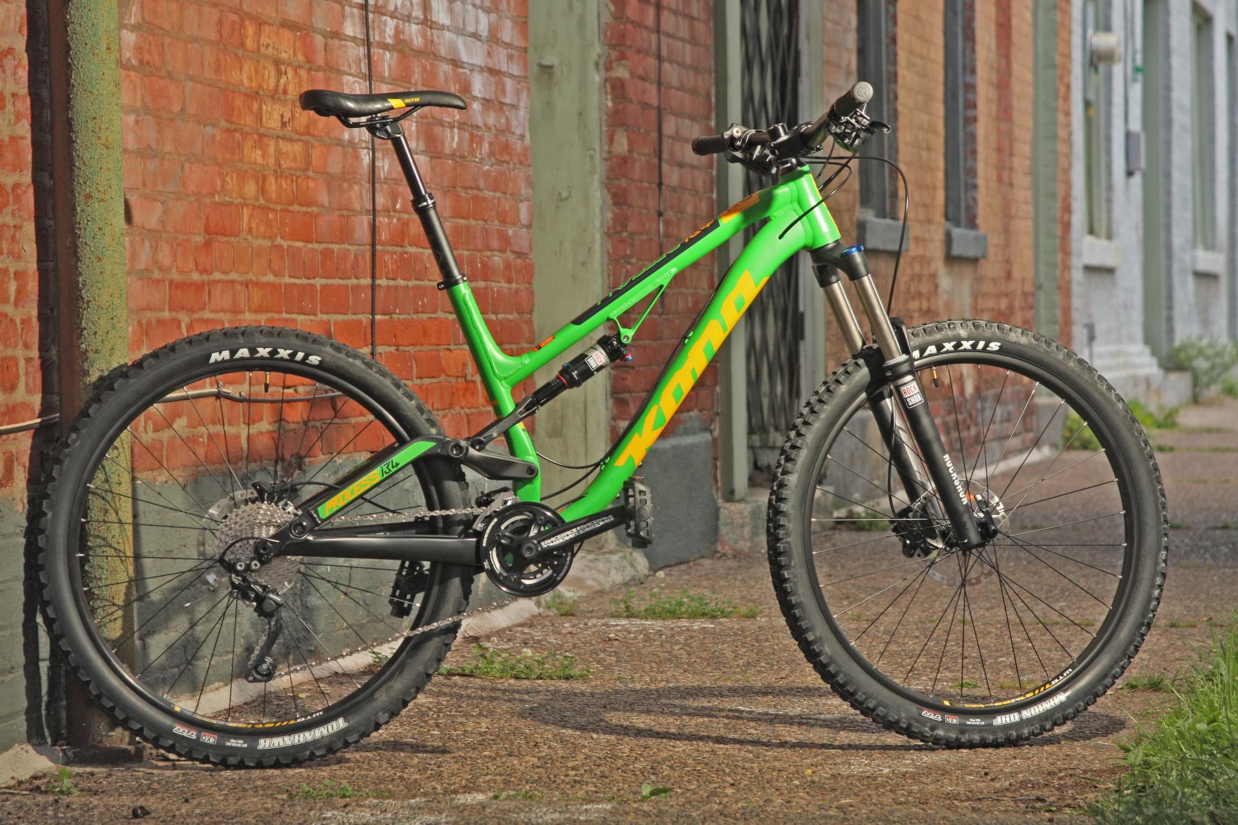 Kona Process 153: A vibrant green Kona mountain bike leaning against a red brick wall, showcasing its sturdy tires and suspension system. The bike is positioned on a gravel path, with faded brick and stone buildings visible in the background.