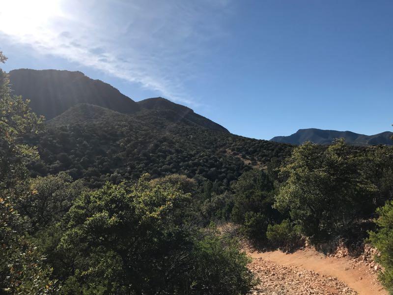 A scenic view of rolling hills and mountains, with lush greenery and a clear blue sky. Sunlight shines over the landscape, highlighting the natural beauty of the terrain. A dirt path winds through the foreground, inviting exploration. Brown Canyon mountain bike trail.