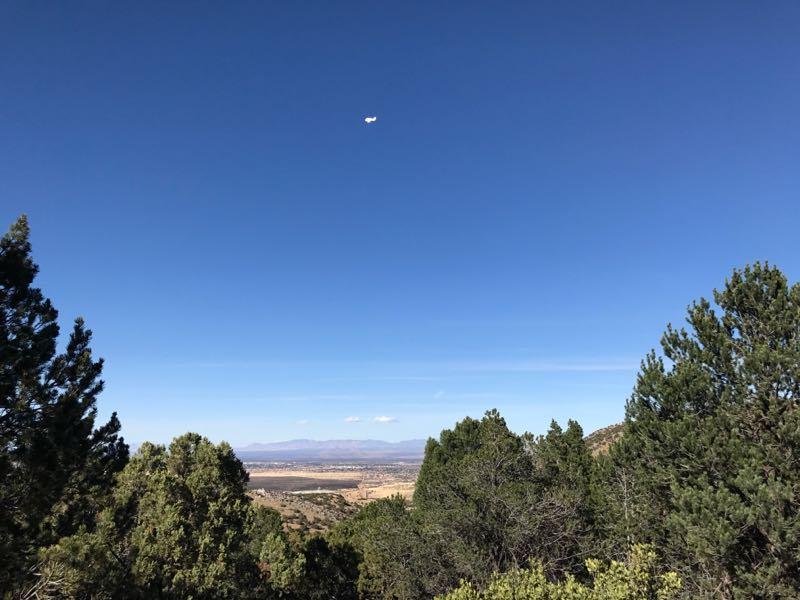 A clear blue sky with a few wispy clouds, featuring a small airplane flying above a scenic landscape. In the foreground, there are green pine trees, while the background showcases rolling hills and valleys under bright sunlight. Brown Canyon mountain bike trail.