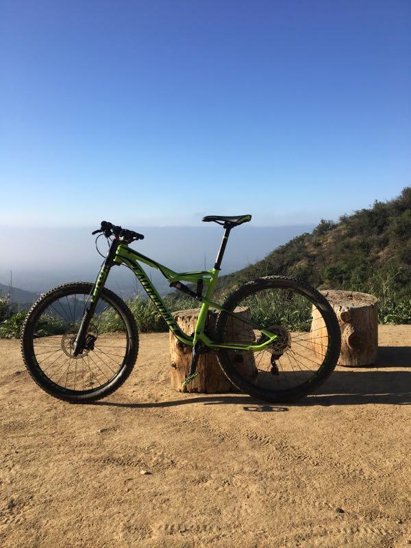 A green mountain bike stands next to two wooden stumps on a dirt path, with a scenic view of hills in the background under a clear blue sky. El Prieto mountain bike trail.
