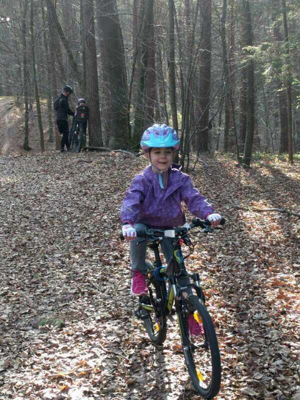 A young girl in a purple jacket and blue helmet rides a bicycle on a leaf-covered path in a forest. In the background, two figures stand next to their bikes among the trees. Sunlight filters through the branches, creating a warm atmosphere. Technikparcours mountain bike trail.