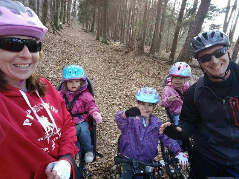 A family enjoying a bike ride in a wooded area. The group includes an adult wearing sunglasses and a pink helmet, along with three young children in pink and blue helmets, seated in bike trailers. They are smiling and posing for a selfie on a leaf-covered path surrounded by trees. Technikparcours mountain bike trail.