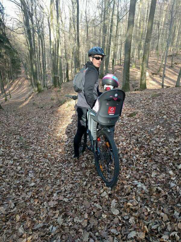 A person wearing a helmet and sunglasses stands beside a mountain bike on a wooded trail covered with fallen leaves, holding the bike while looking back. A child in a safety seat is positioned on the back of the bike, also wearing a helmet. The surroundings include tall trees with bare branches, typical of early spring. Technikparcours mountain bike trail.