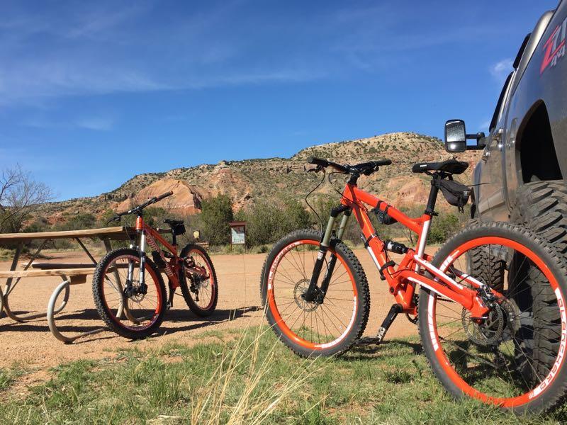 Two mountain bikes, one orange and one green with orange accents, are leaning against a large pickup truck. In the background, red rock formations rise against a clear blue sky, and a picnic table is visible nearby. The scene is set in an outdoor area, surrounded by grass and sparse vegetation. Palo Duro Canyon mountain bike trail.
