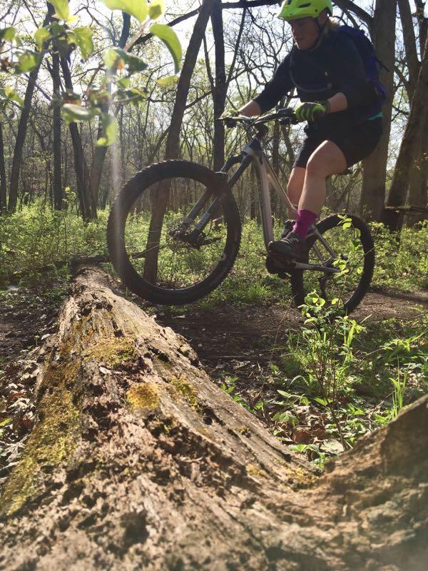 A person riding a mountain bike on a forest trail, balancing on a fallen log surrounded by lush green foliage. The cyclist is wearing a bright yellow helmet and purple socks, demonstrating an action shot in a sunny outdoor setting. Shawnee Mission Park mountain bike trail.