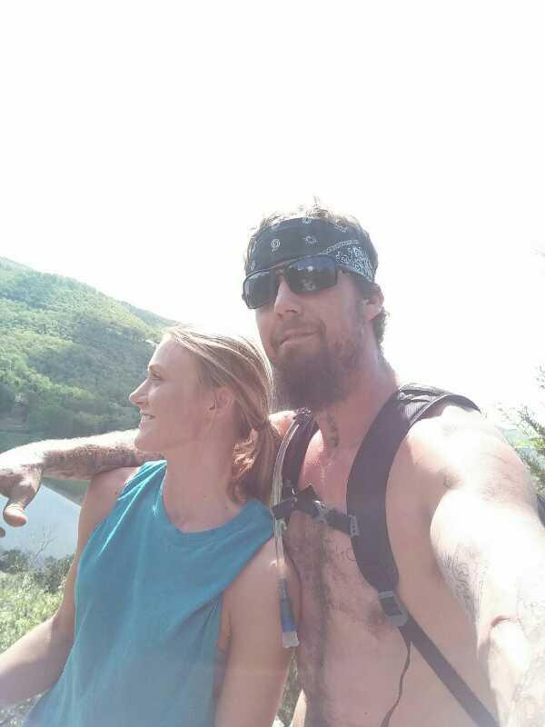 A man and a woman pose for a selfie outdoors, with green hills and water visible in the background. The man is shirtless, wearing sunglasses and a bandana, while the woman is wearing a tank top and smiling at him. The sunlight brightens the scene, creating a cheerful and adventurous atmosphere. Syllamo Trails mountain bike trail.
