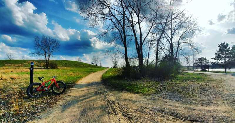 A scenic outdoor path splits between grassy areas, with a red bicycle resting against a post on the left. Leafless trees line the path, and a bright blue sky with fluffy clouds overhead adds to the tranquil atmosphere. Island Lake mountain bike trail.