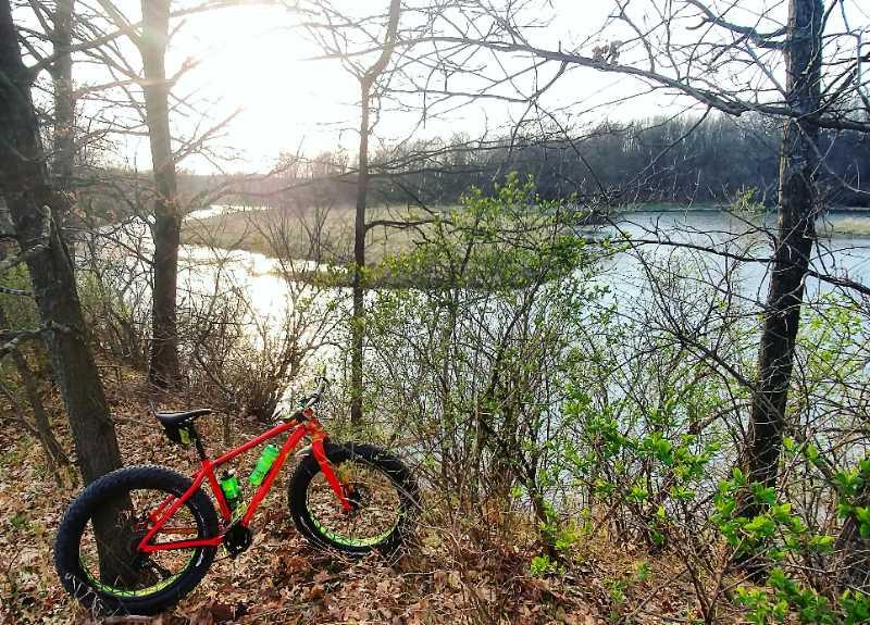 A vibrant red fat bike resting on the ground near a serene riverbank, surrounded by sparse trees and budding greenery, with a soft evening glow from the setting sun illuminating the scene. Island Lake mountain bike trail.