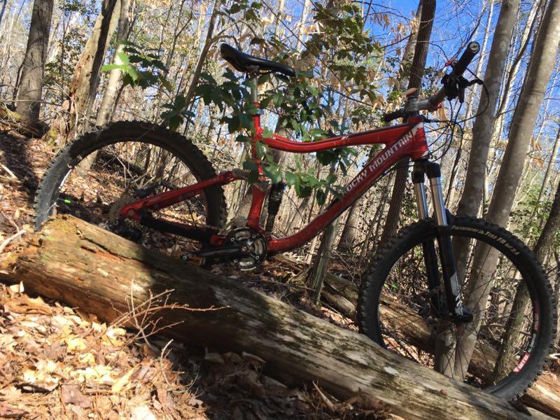A red mountain bike leaning against a fallen log in a wooded area, with trees and foliage in the background. Sunlight filters through the branches, highlighting the bike and surrounding nature. Mountain Laurel Trails mountain bike trail.