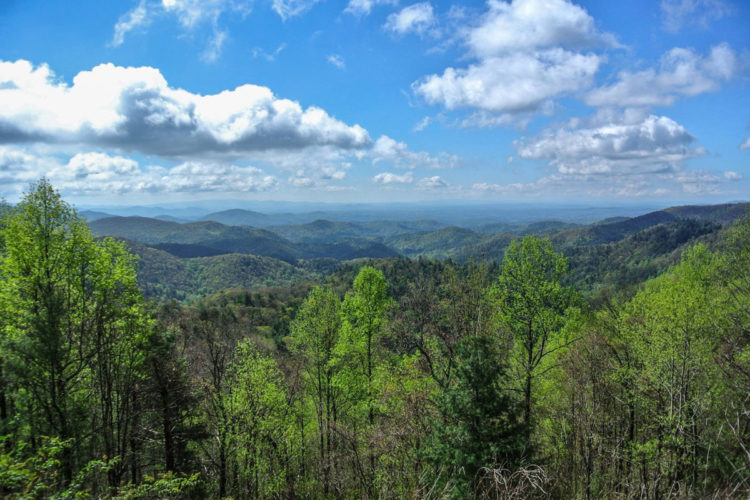 A panoramic view of lush green mountains under a bright blue sky with scattered clouds. The landscape features rolling hills, vibrant trees, and various shades of green, creating a serene and picturesque natural scene.