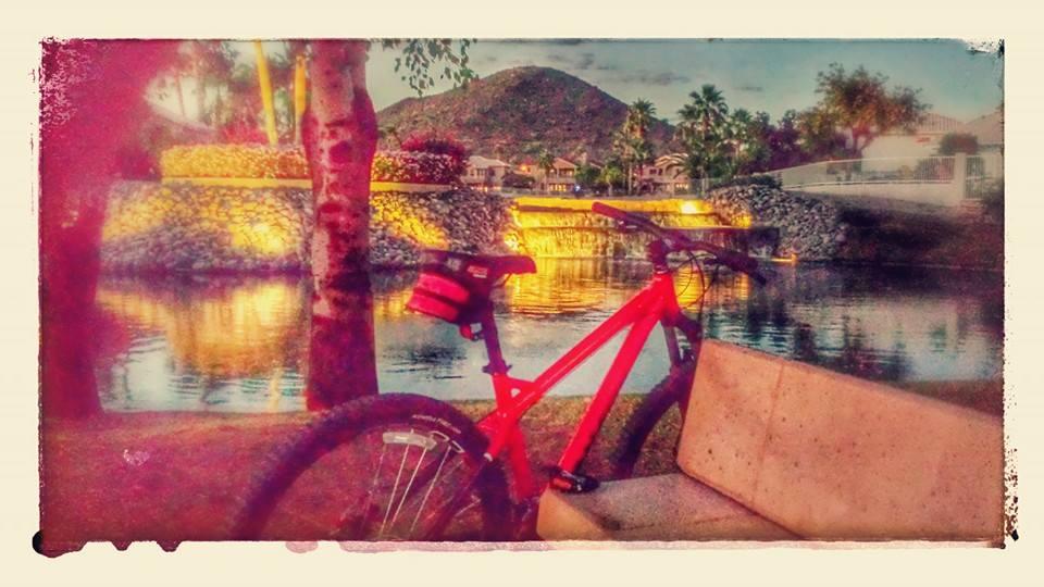 Raleigh Tokul: A red mountain bike resting on a concrete bench near a serene pond, surrounded by palm trees and illuminated by soft lights. In the background, a gentle waterfall flows down a stone wall, with a mountain rising in the distance under a twilight sky.