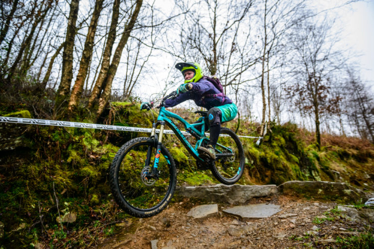 A mountain biker in a green helmet and purple jacket jumps off a rocky terrain, navigating a forested path with trees in the background. The bike features a turquoise frame and sturdy tires, indicating an adventurous ride through a mossy landscape.