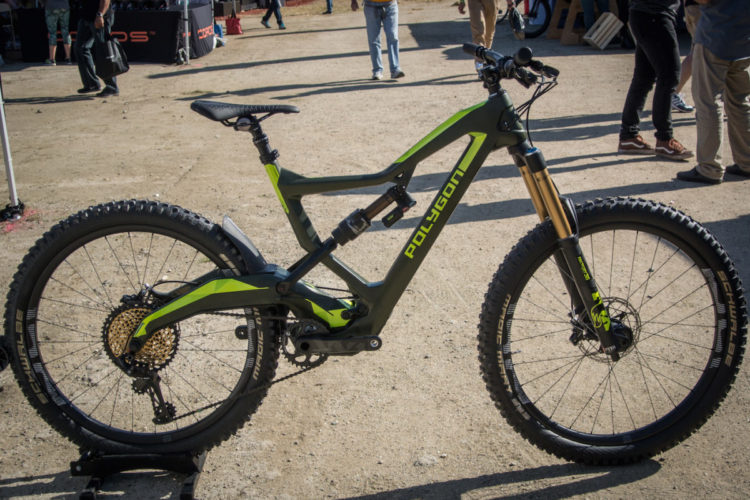 A black and green Polygon mountain bike displayed on a gravel surface, showcasing its sleek design and features, including a full suspension system and thick tires. In the background, a blurred crowd of people and tents can be seen, suggesting a biking event or expo.