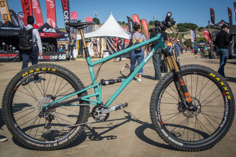 A mountain bike with a teal frame and black Maxxis tires, displayed at an outdoor event with colorful flags and tents in the background. The bike features a front suspension fork and a rear shock. People are walking in the background, engaging with the event.