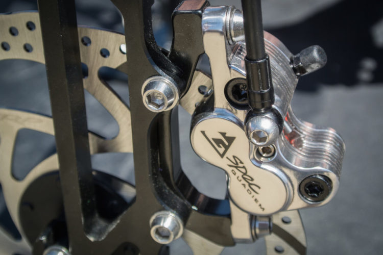 Close-up of a bicycle disc brake system, featuring a metallic brake caliper and rotor. The image shows intricate mechanical details, including bolts and mounting components, highlighting the brake's design and functionality.