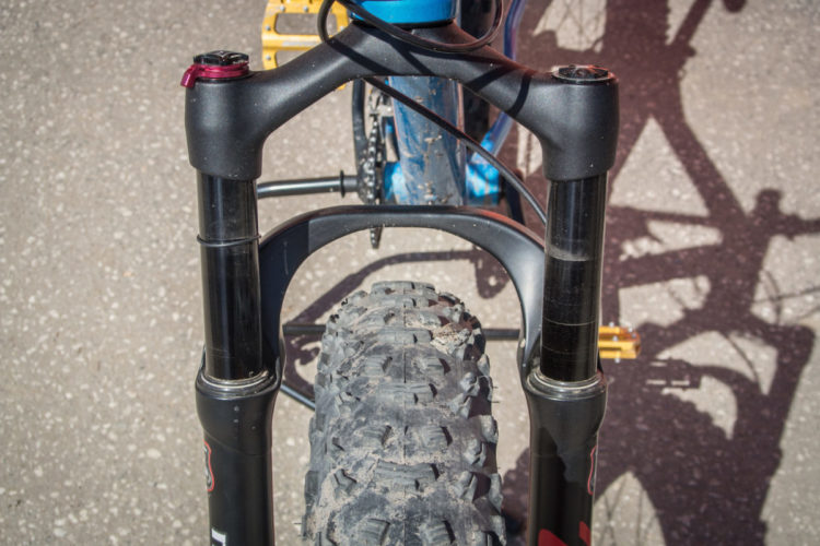 Close-up view of a mountain bike front fork, showing the tire and suspension components. The image highlights the rugged tread of the tire and the sleek design of the fork, set against a textured gravel surface.