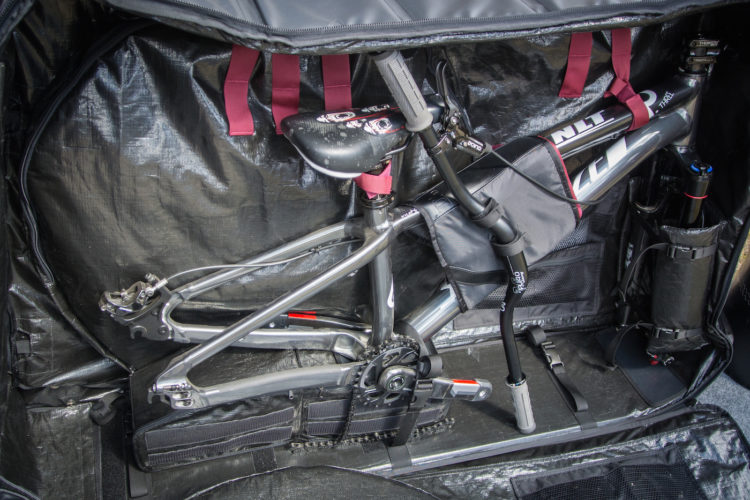 A disassembled bicycle secured inside a protective travel bag, featuring a black interior with organizational straps and pockets for accessories. The bike's frame is prominently displayed, with pedals and handlebars positioned for transport.