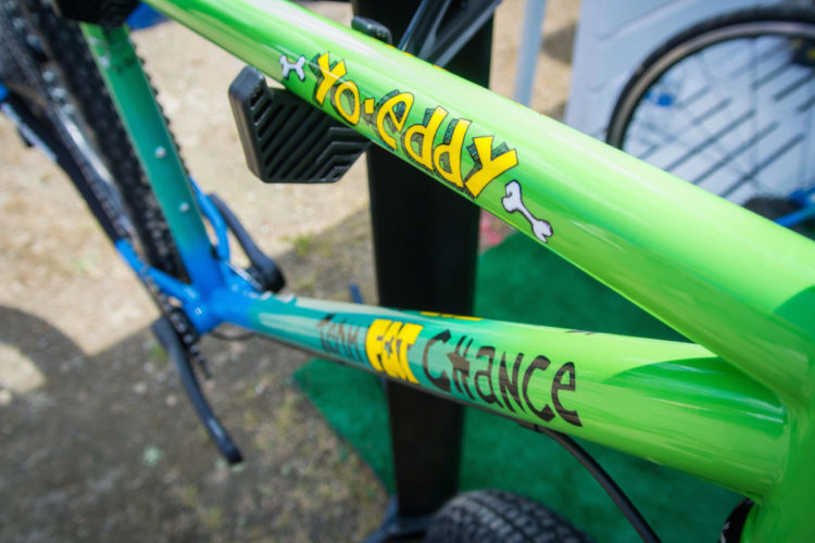 Close-up view of a colorful bicycle frame featuring the text "Yo Eddy" in bright yellow and "Fat Chance" in bold brown lettering, alongside playful bone graphics. The frame displays a vibrant green and blue color scheme, set against a blurred outdoor background.