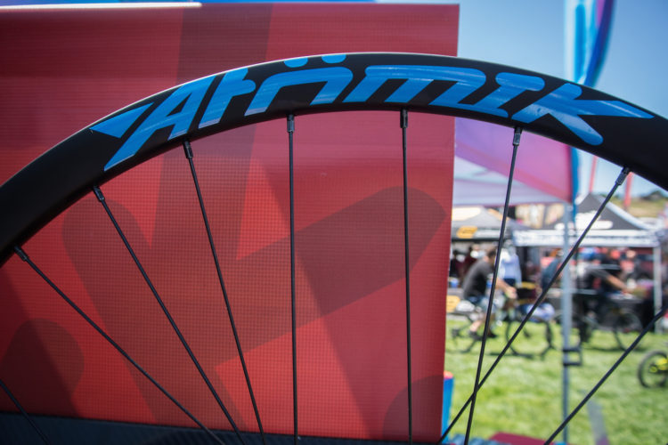 A close-up view of a bicycle wheel rim featuring the logo "Atörmik" in blue, set against a blurred background of a vibrant red display. Visible spokes and the structure of the wheel are in focus, while outdoor event tents and people with bicycles are slightly out of focus in the background.