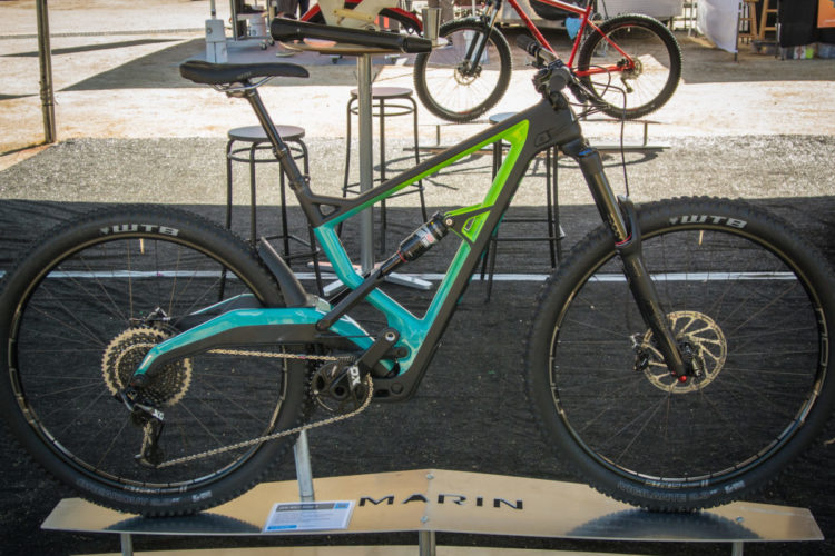 Alt text: A modern mountain bike displayed on a stand, featuring a teal and green frame with thick tires. The bike has a dual-suspension design, visible gear components, and is set in a venue with a black backdrop and other bicycles in the background.