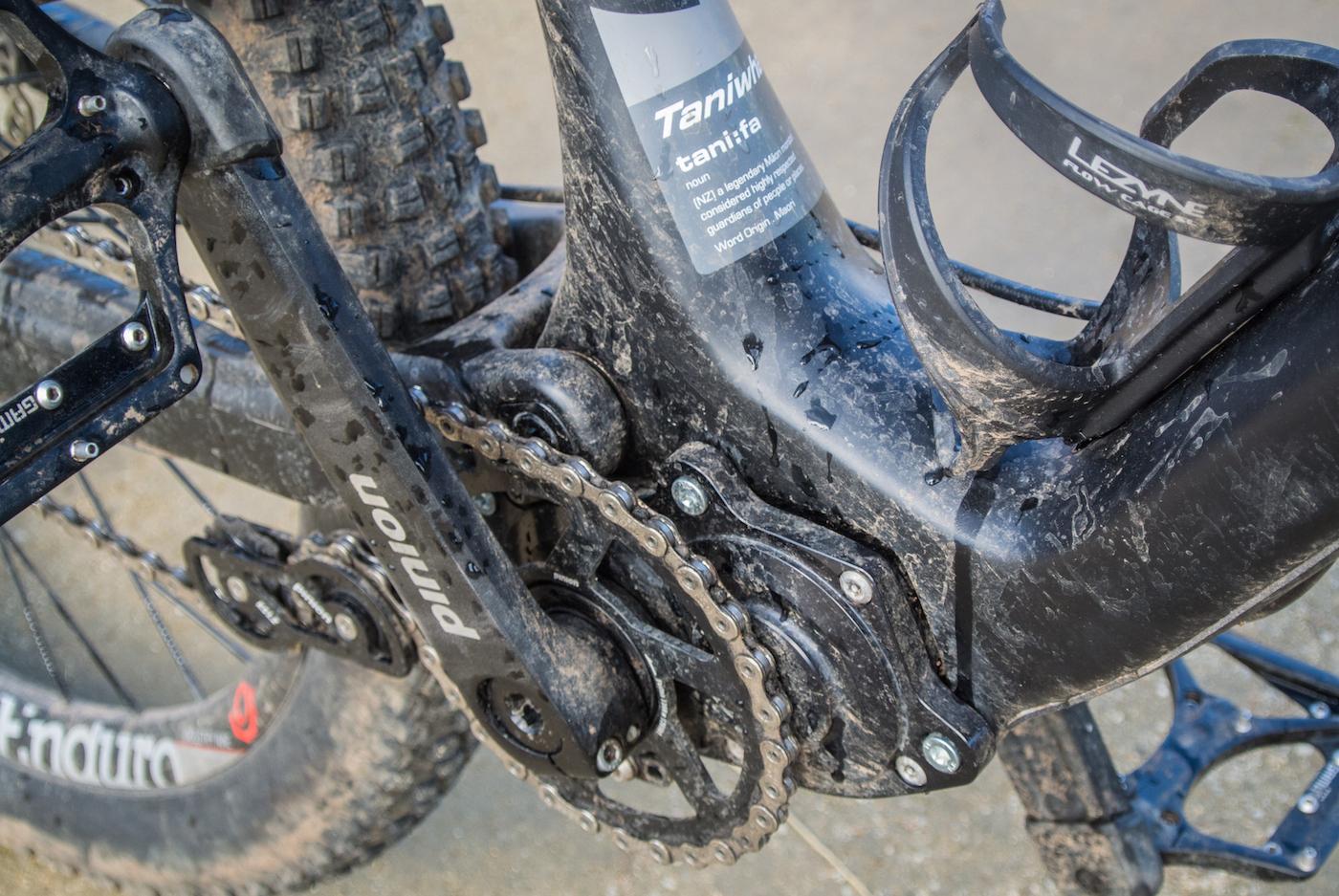 Zerode Taniwha: Close-up view of a mountain bike's drivetrain, showing the crankset, chain, and gearing mechanism. The components are covered in dirt and mud, indicating recent use on rugged terrain. A water bottle holder is also visible on the frame.