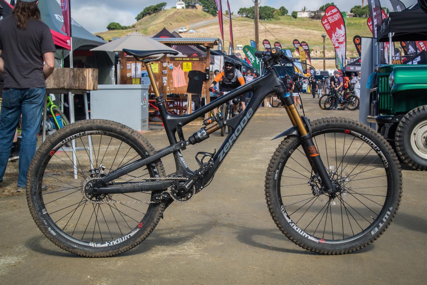 Zerode Taniwha: A black mountain bike with a sleek frame, featuring a gold suspension fork and rugged tires, is positioned on a dirt surface. The background includes a busy outdoor event setup with various tents, bicycles, and people.
