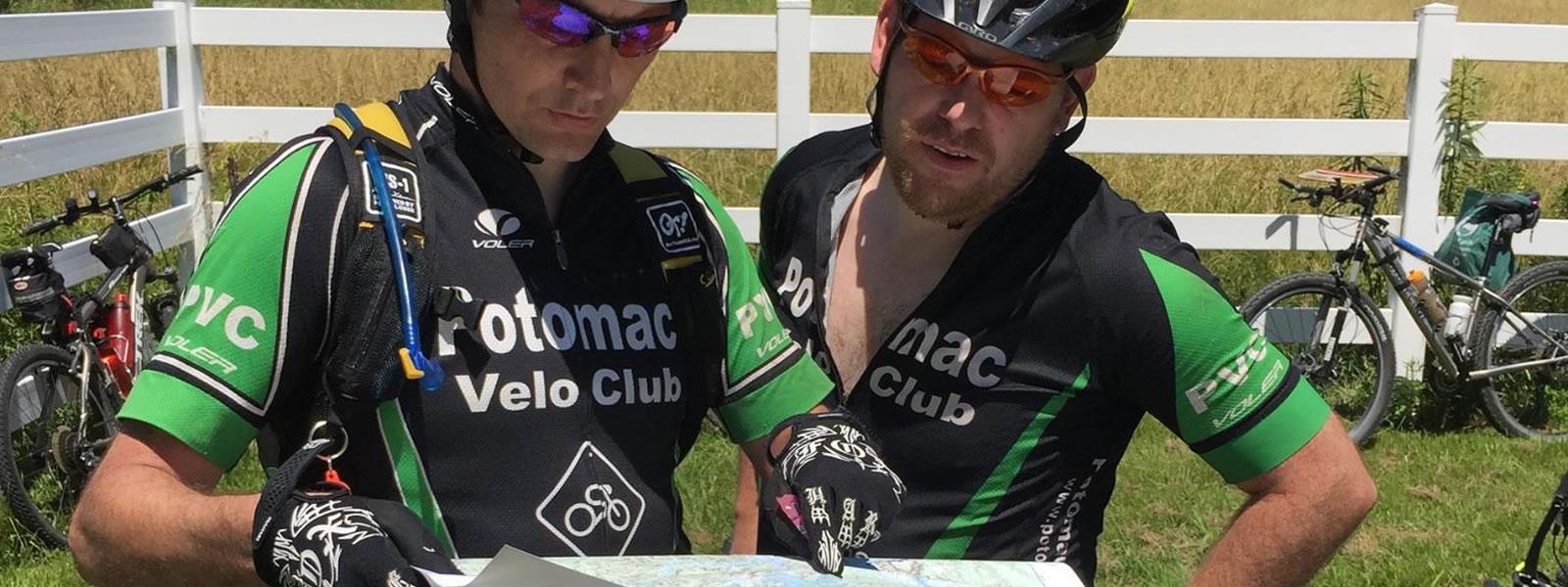 Two cyclists in matching jerseys from the Potomac Velo Club are closely examining a map outdoors. They are surrounded by green grass and a white fence, with bicycles parked in the background. One cyclist is wearing sunglasses and gripping the map, while the other leans in to get a better view. Both appear focused on their navigation.